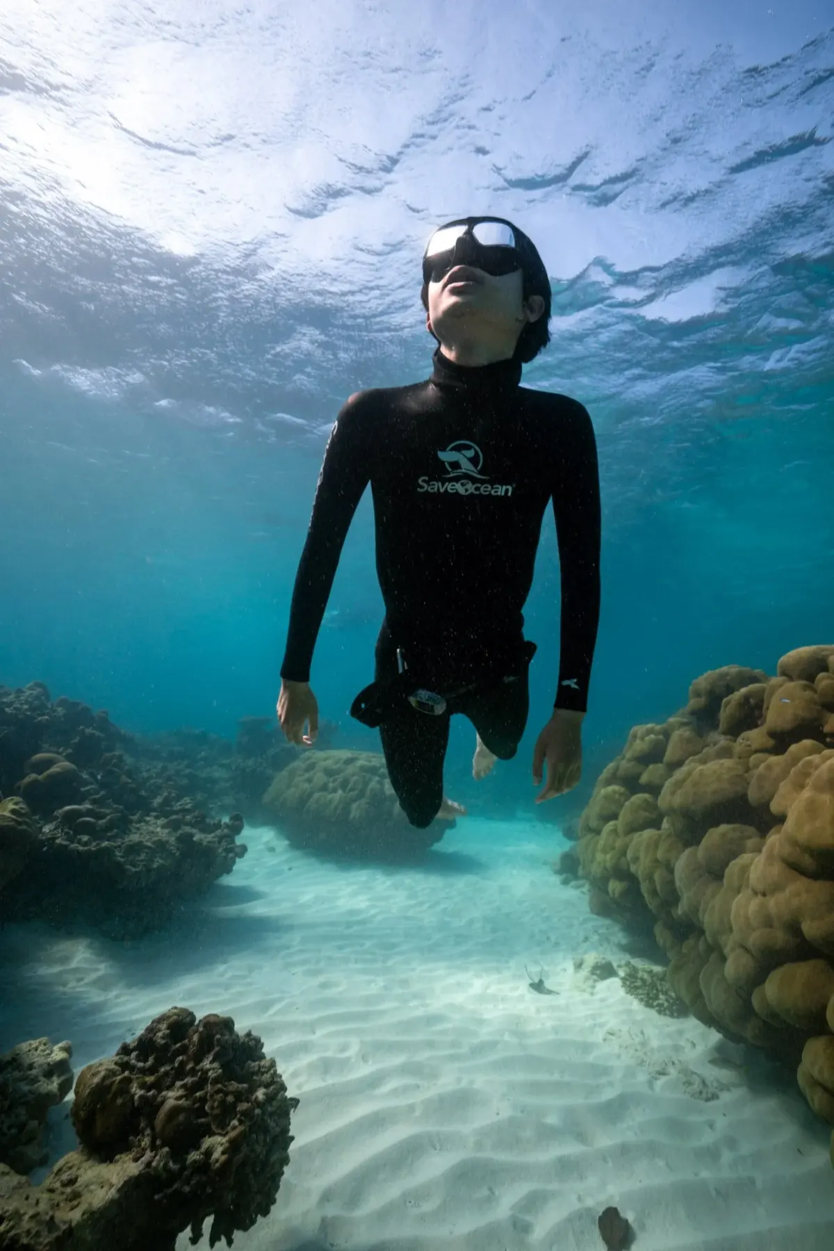 Freediver in a wetsuit hovering above the white sand between coral heads off Koh Tao