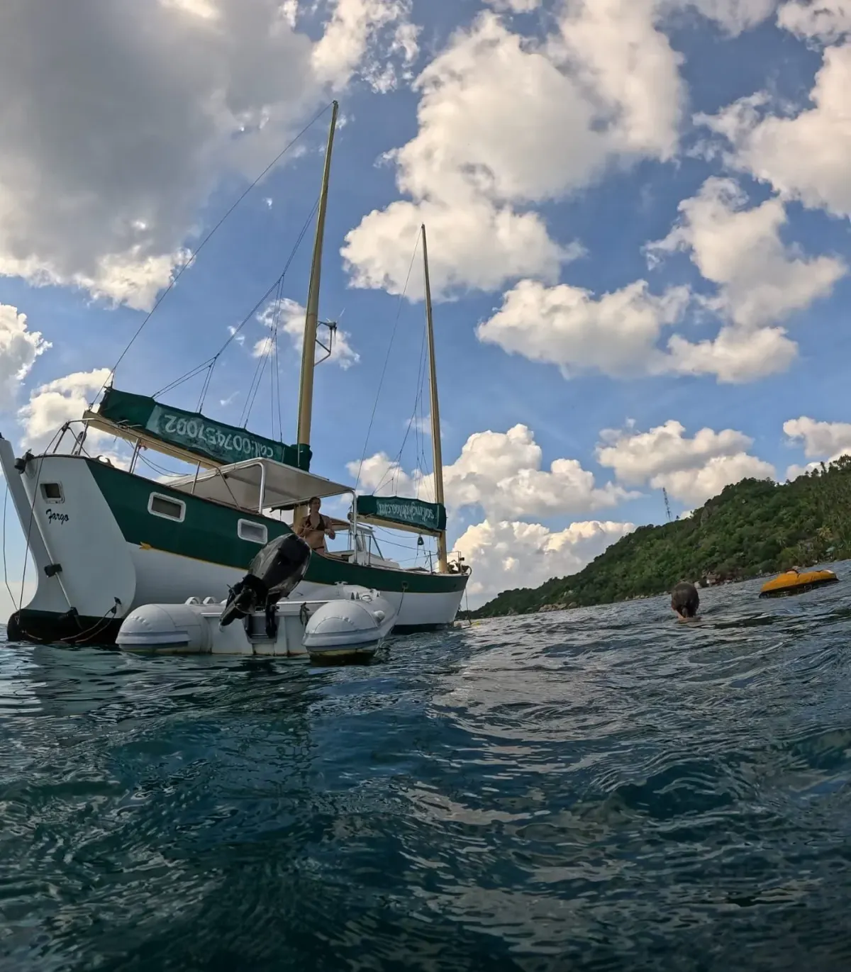 SV Freedom Fargo seen from the water, with the boat dog standing on the stern and a swimmer alongside