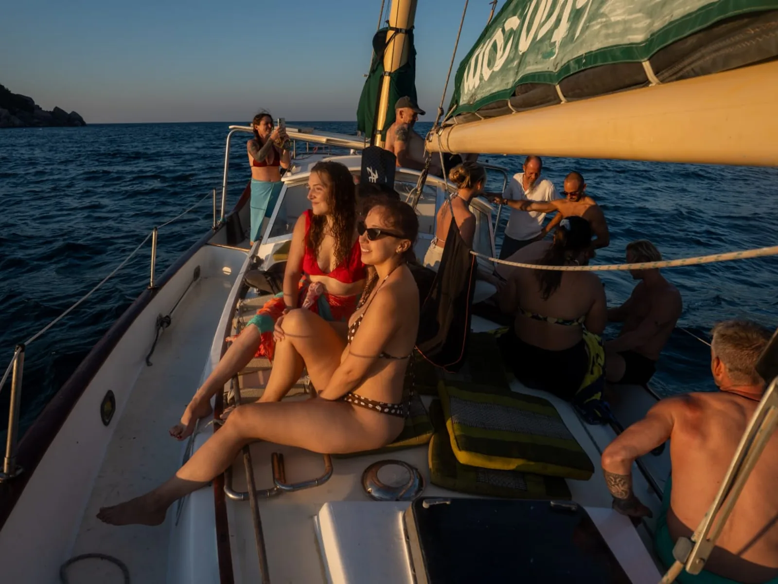 Guests smiling and relaxing along the deck of SV Freedom Fargo in golden evening light during a sunset sail off Koh Tao